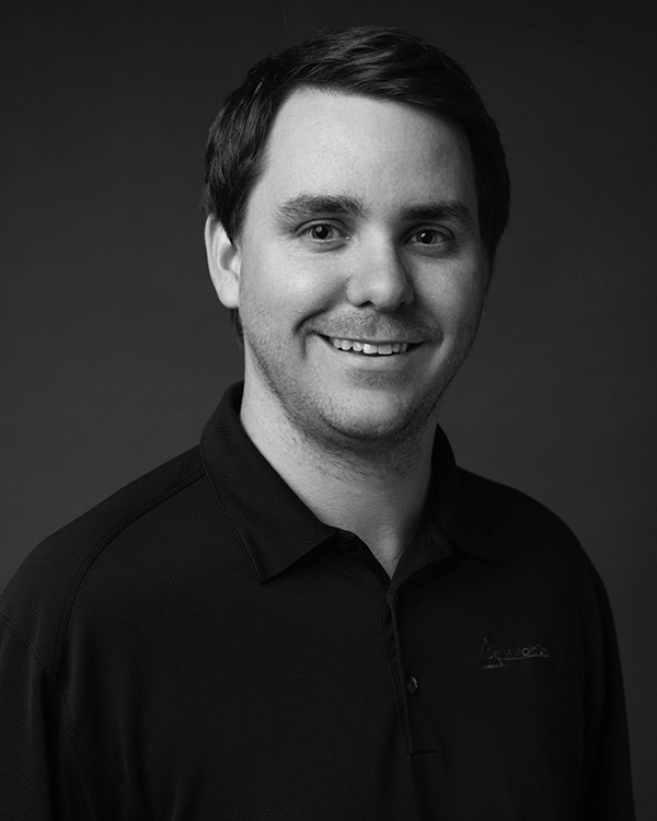 A man with short dark hair wearing a dark collared shirt smiles while facing the camera, against a plain dark background.