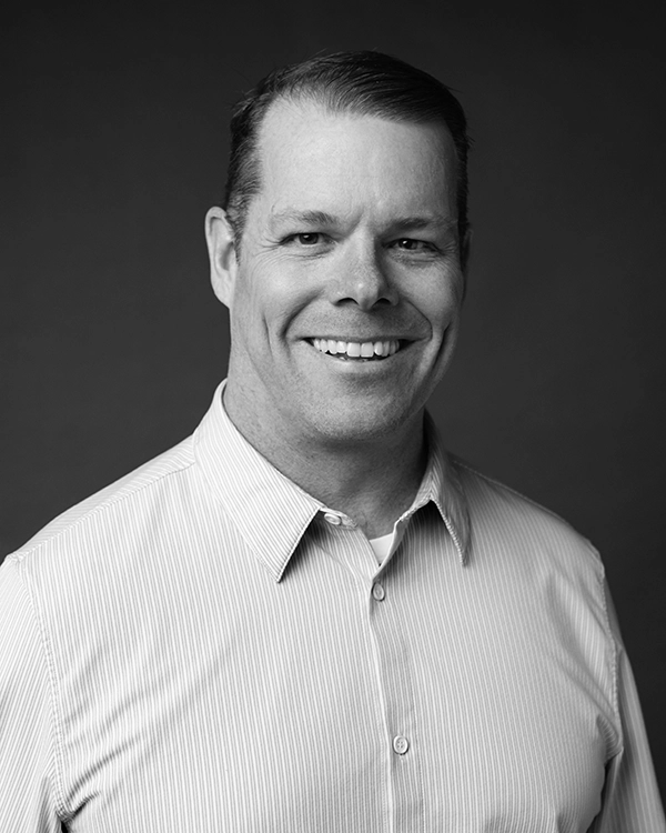 Smiling man with short hair wearing a light-colored, striped button-up shirt, posed against a plain dark background.