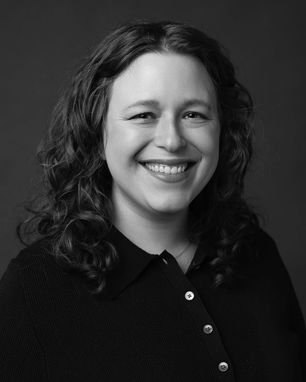 A person with wavy hair and a collared shirt smiles at the camera against a plain, dark background. The image is in black and white.
