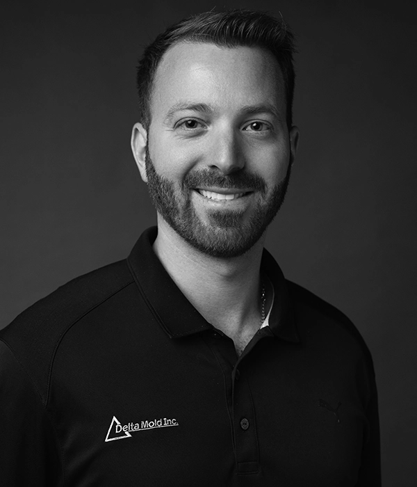 A man with short hair and a beard, wearing a black polo shirt with the Delta Mold Inc. logo, poses and smiles in front of a plain dark background.