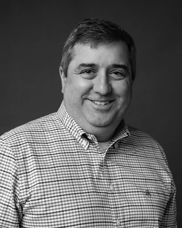 A man with short dark hair, wearing a checkered button-up shirt, smiling and standing in front of a plain dark background.