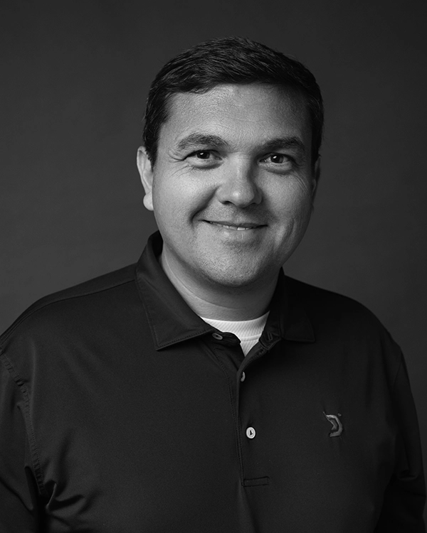 Man in a dark polo shirt smiles at the camera against a plain, dark background in a black and white portrait.