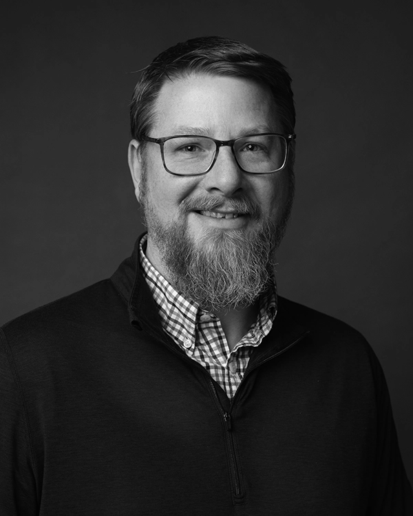 Black and white portrait of a man with glasses, a beard, and a mustache, wearing a checkered shirt under a zip-up sweater, looking at the camera and smiling slightly.