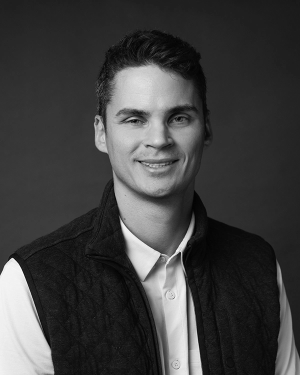 A man with short dark hair, wearing a collared shirt and a quilted vest, is smiling at the camera against a plain dark background.